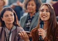 woman with a microphone speaking within a crowd of other seated people